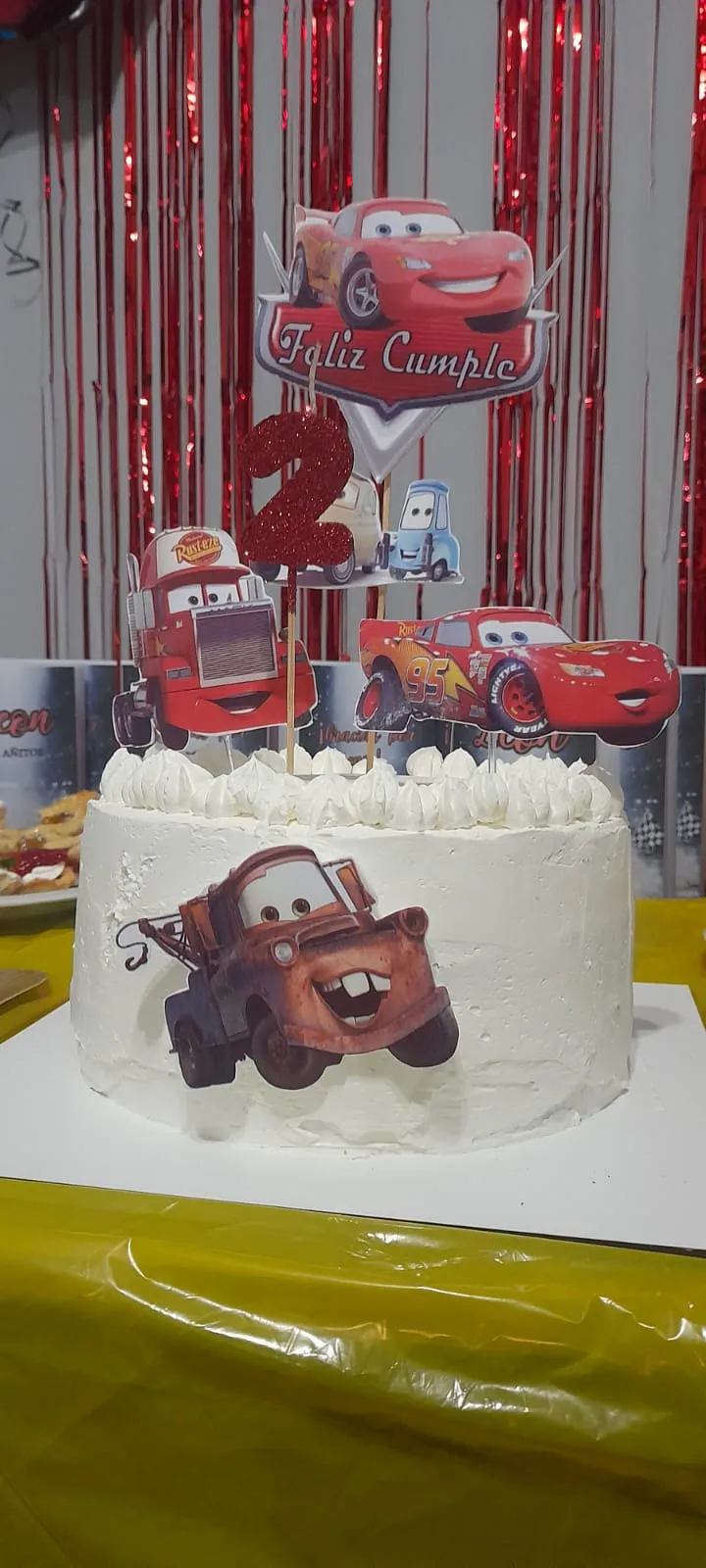 Torta blanca con borde de merengue, figuras de autos de dibujitos y topper de «Feliz Cumple», servida en la mesa del cumpleaños.